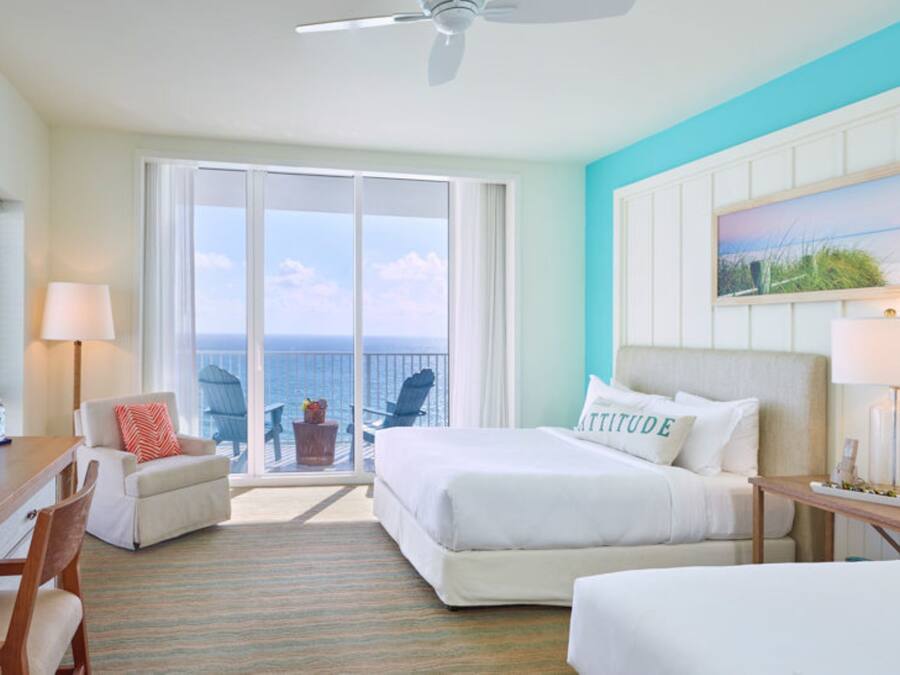 A hotel room with a bed, a desk, a chair, and a view of the ocean through large windows.