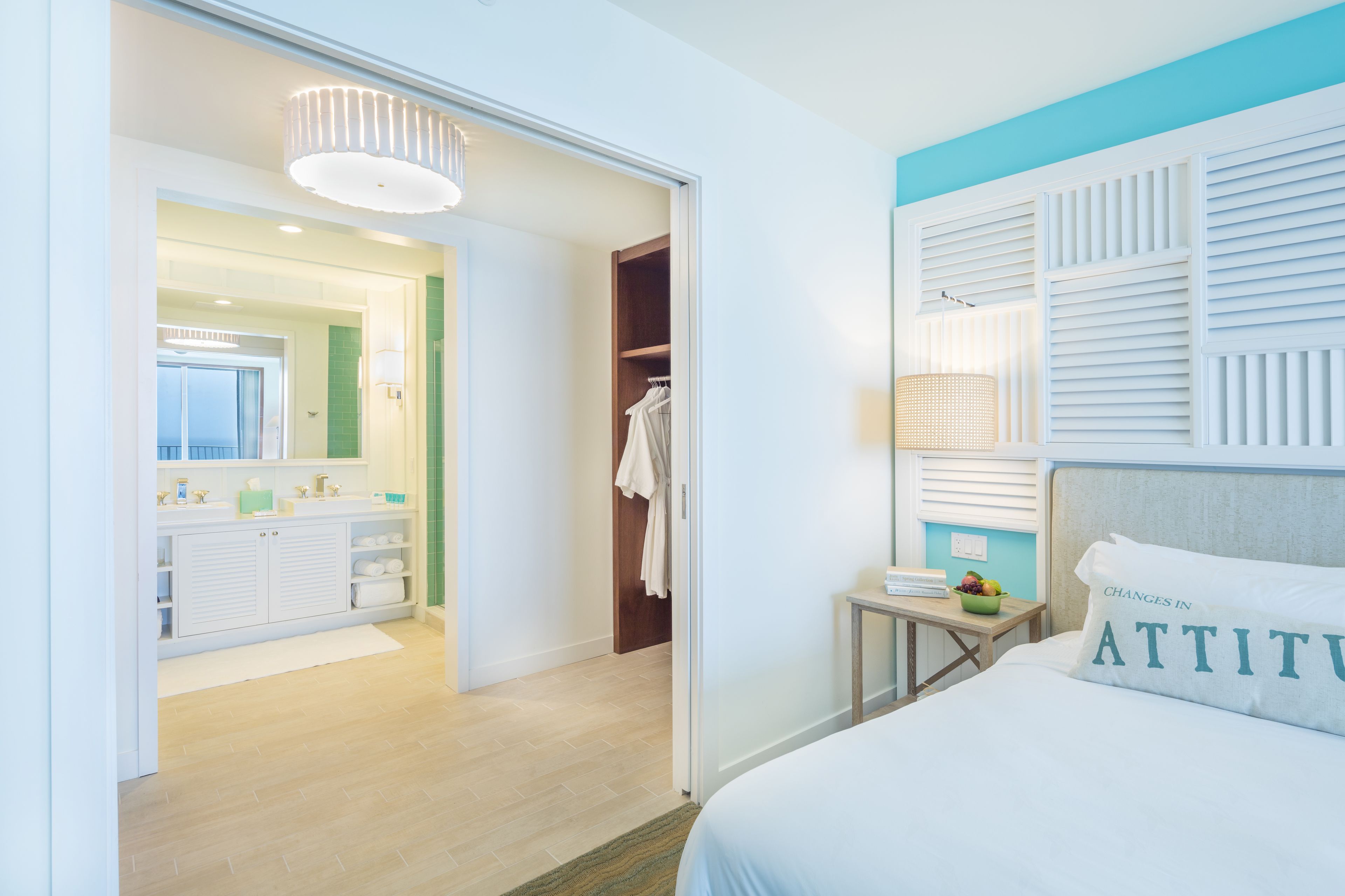 Habitación de hotel con cama, mesita de noche con lámpara, espejo, baño con lavamanos y toallero, y armario con ropa colgada.