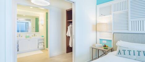 Habitación de hotel con cama, mesita de noche con lámpara, espejo, baño con lavamanos y toallero, y armario con ropa colgada.