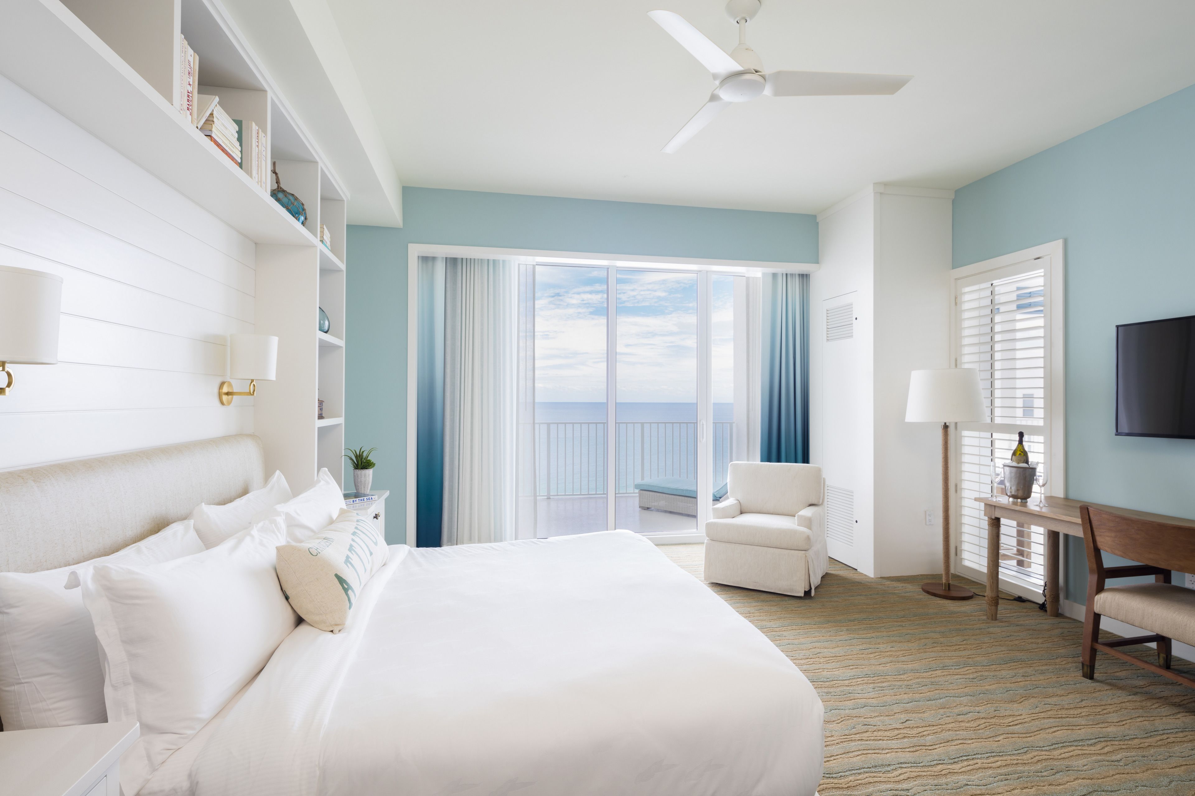 A hotel room with a large bed, a desk, a chair, a TV, and a balcony with a view of the sea.