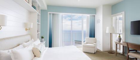 A hotel room with a large bed, a desk, a chair, a TV, and a balcony with a view of the sea.