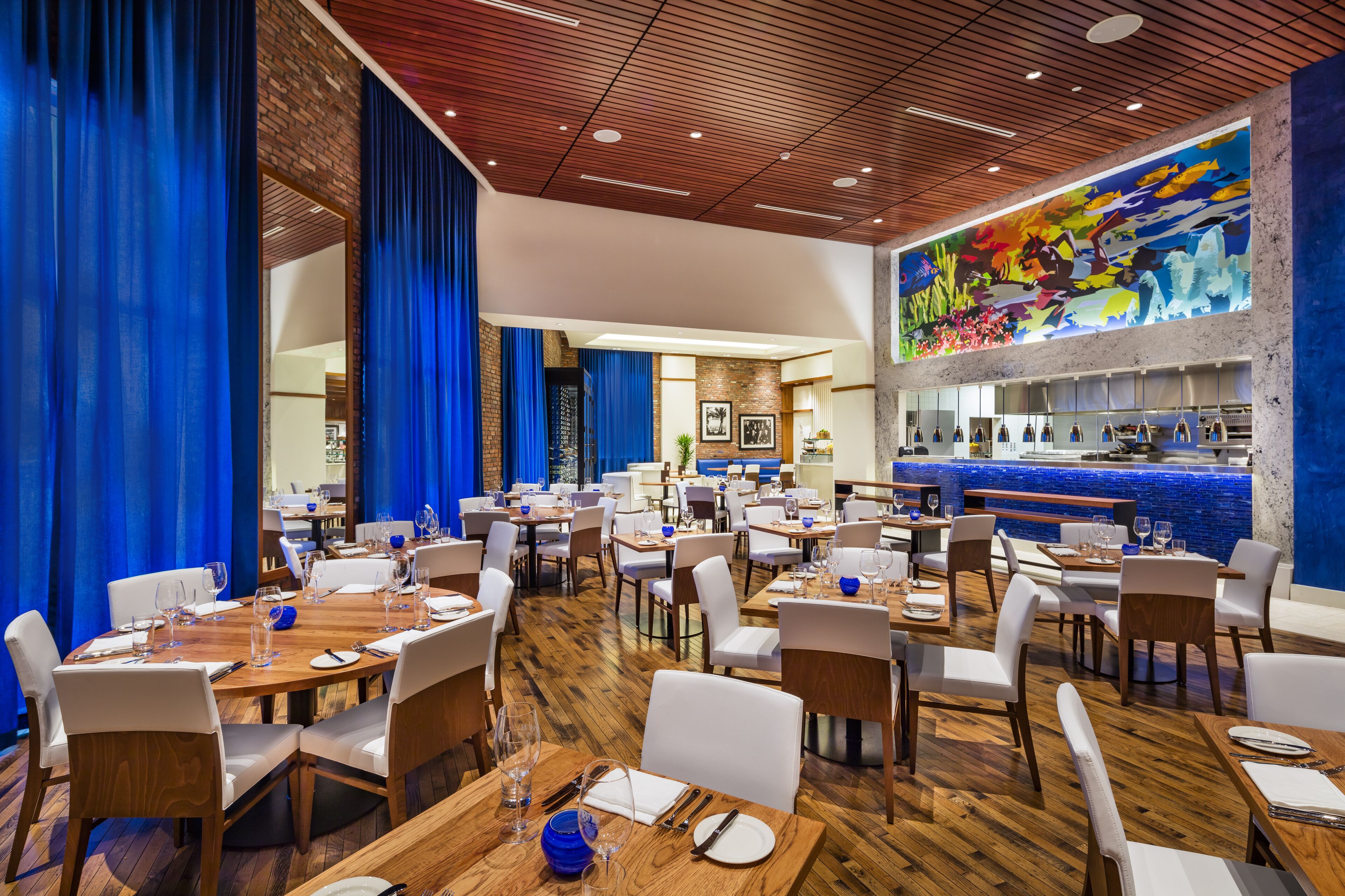 Un restaurante con mesas de madera y sillas blancas, una zona de bar y un mural colorido en la pared.