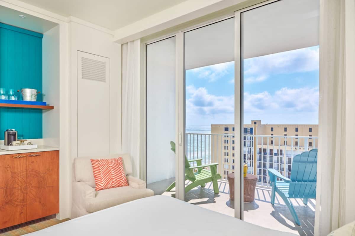 Une chambre d’hôtel comprenant un lit, une chaise, une petite table, un coin cuisine et un balcon avec des chaises et une vue sur la ville.