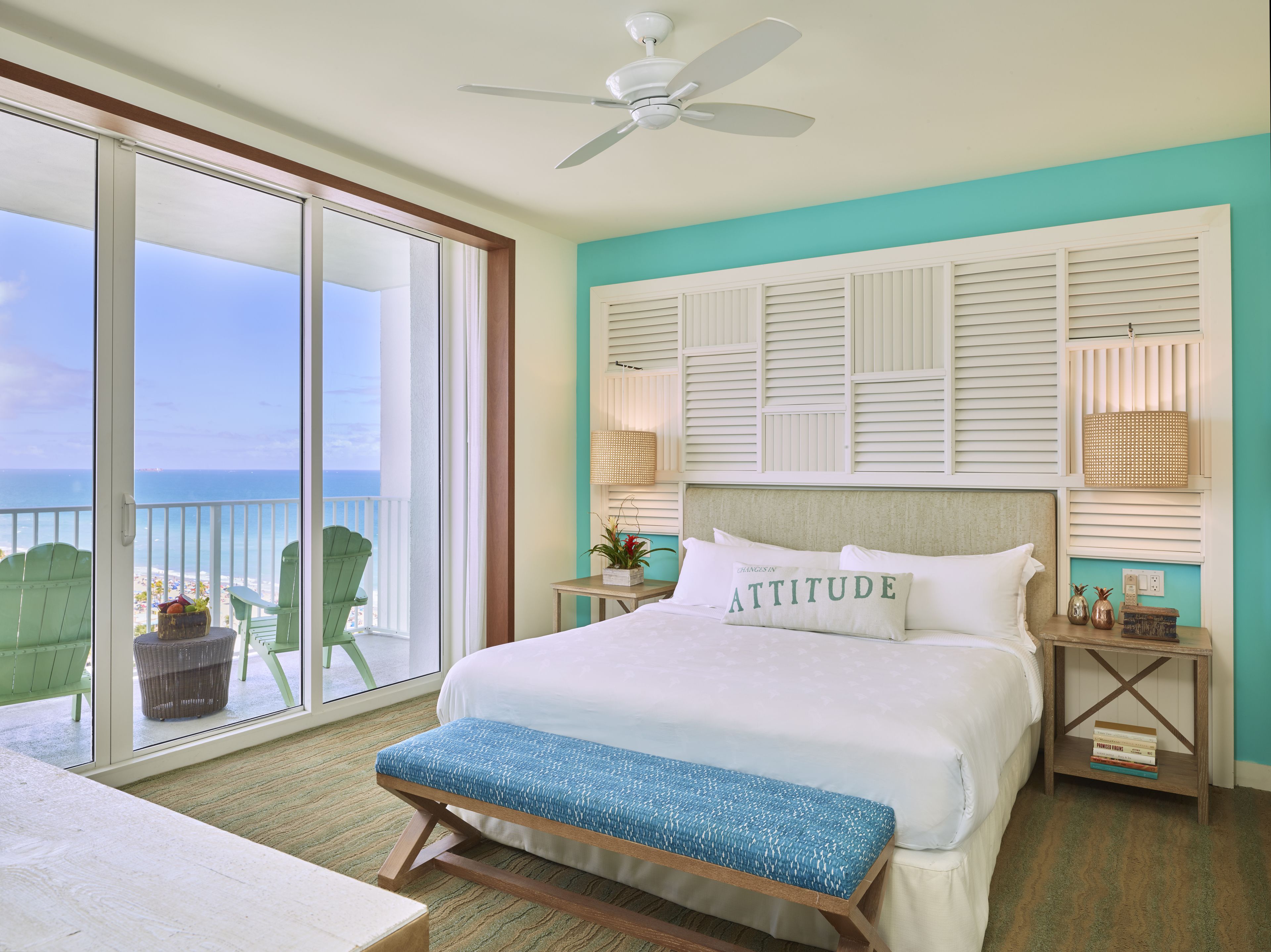 A bedroom with a large bed, a bench, a ceiling fan, a nightstand, a lamp, and a view of the beach.