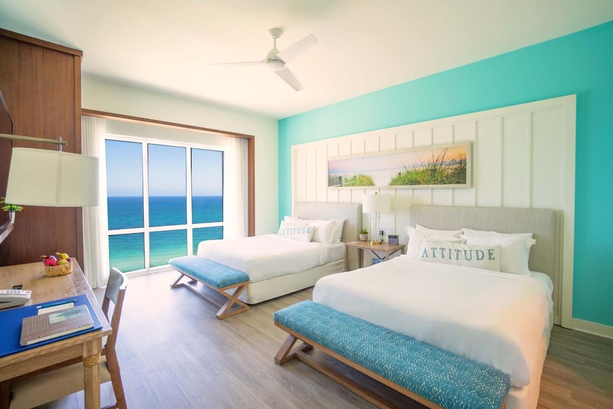 Une chambre d’hôtel avec deux lits, un bureau avec une lampe, une vue sur l’océan et un ventilateur au plafond.