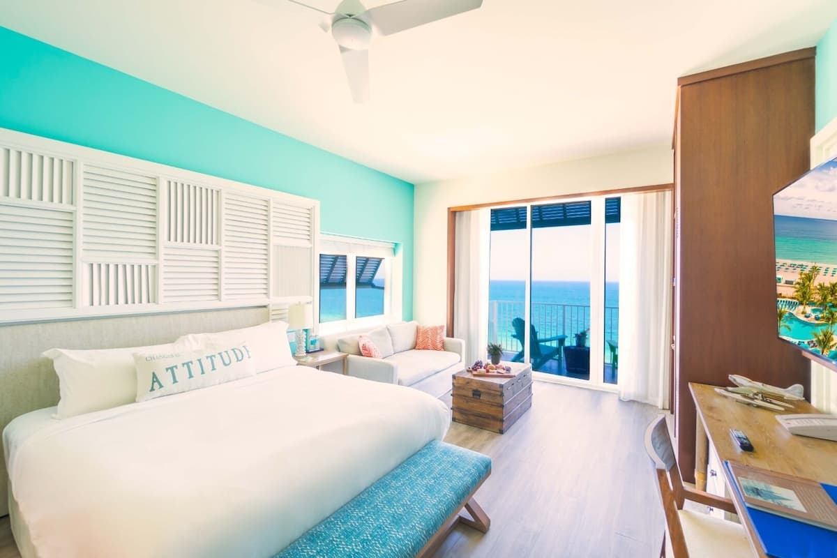 Une chambre d’hôtel dotée d’un grand lit, d’un canapé, d’une commode en bois et d’une table à manger. La chambre offre une vue sur l’océan et dispose d’un balcon aménagé avec du mobilier d’extérieur.