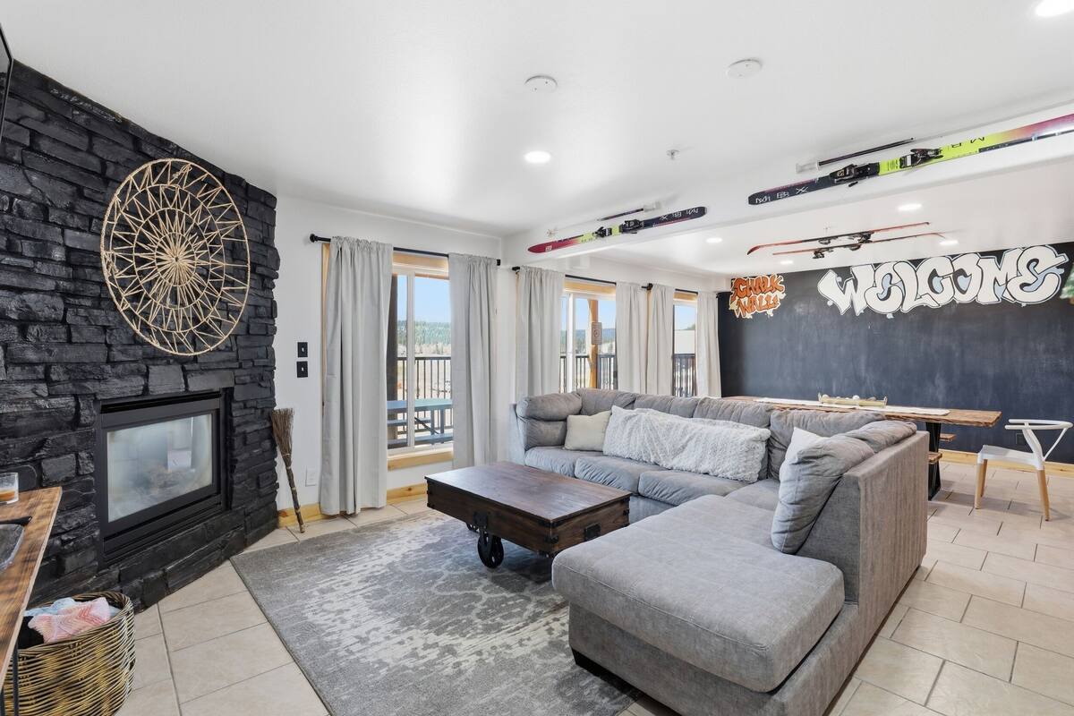 A modern living room with a stone fireplace, a sectional sofa, a coffee table, and ski equipment on the ceiling.