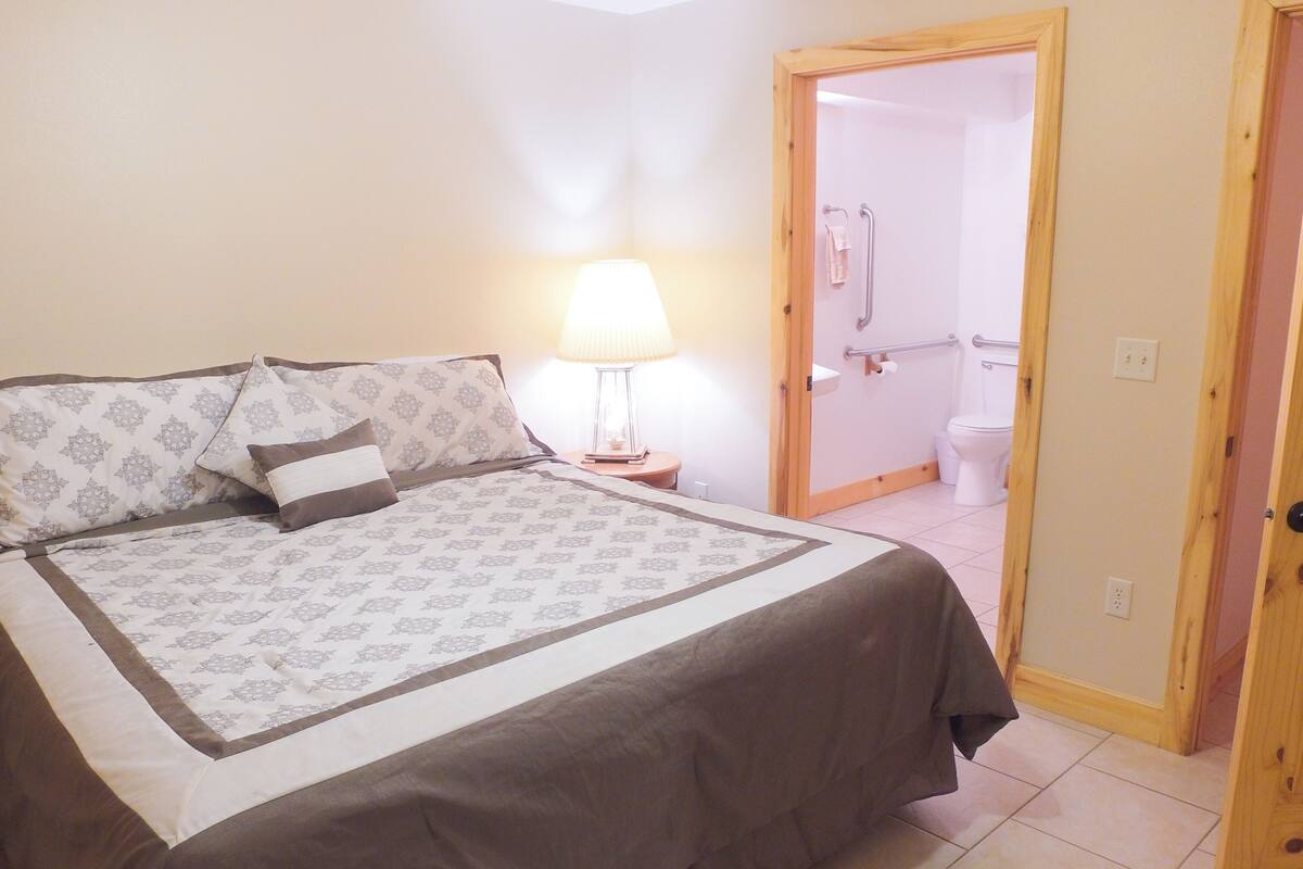 A bedroom with a bed, a bedside lamp, a nightstand, and a bathroom visible through an open door.