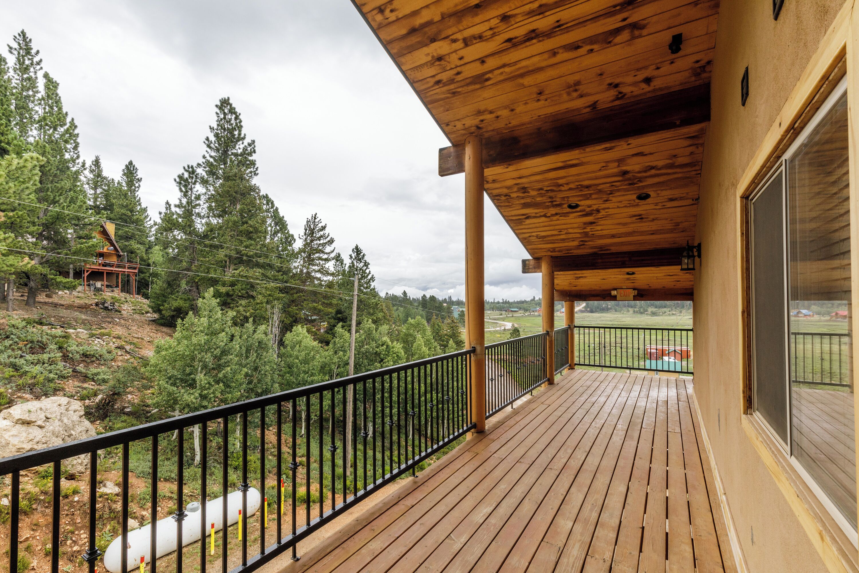 A wooden deck with a black railing, overlooking a forested area with a view of a distant building.