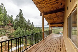 A wooden deck with a black railing, overlooking a forested area with a view of a distant building.