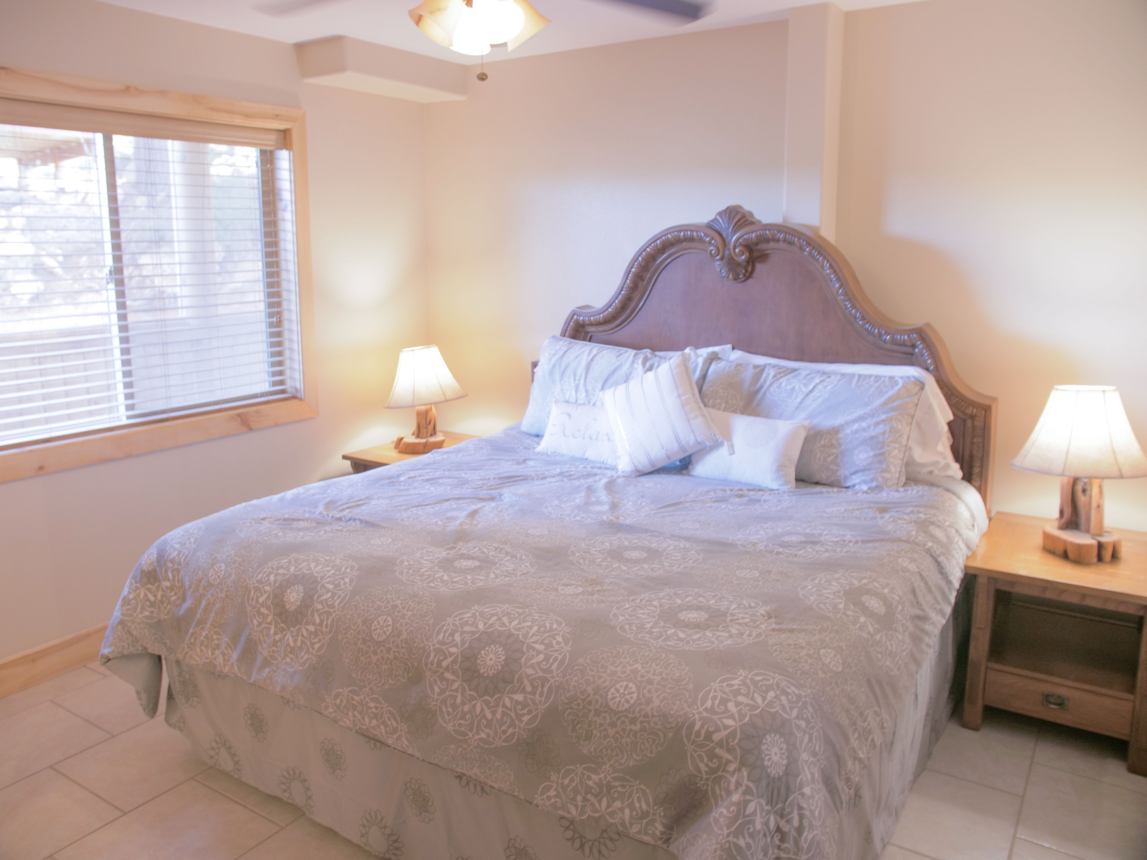 A bedroom with a large bed, two bedside tables, a window with blinds, and a ceiling fan.