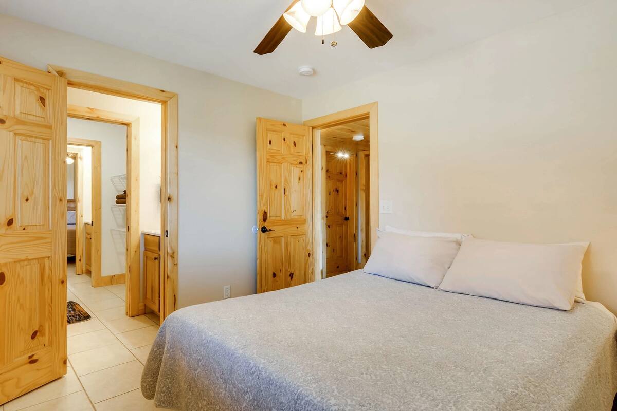 A bedroom with a bed, wooden doors, a ceiling fan, and a visible bathroom in the background.