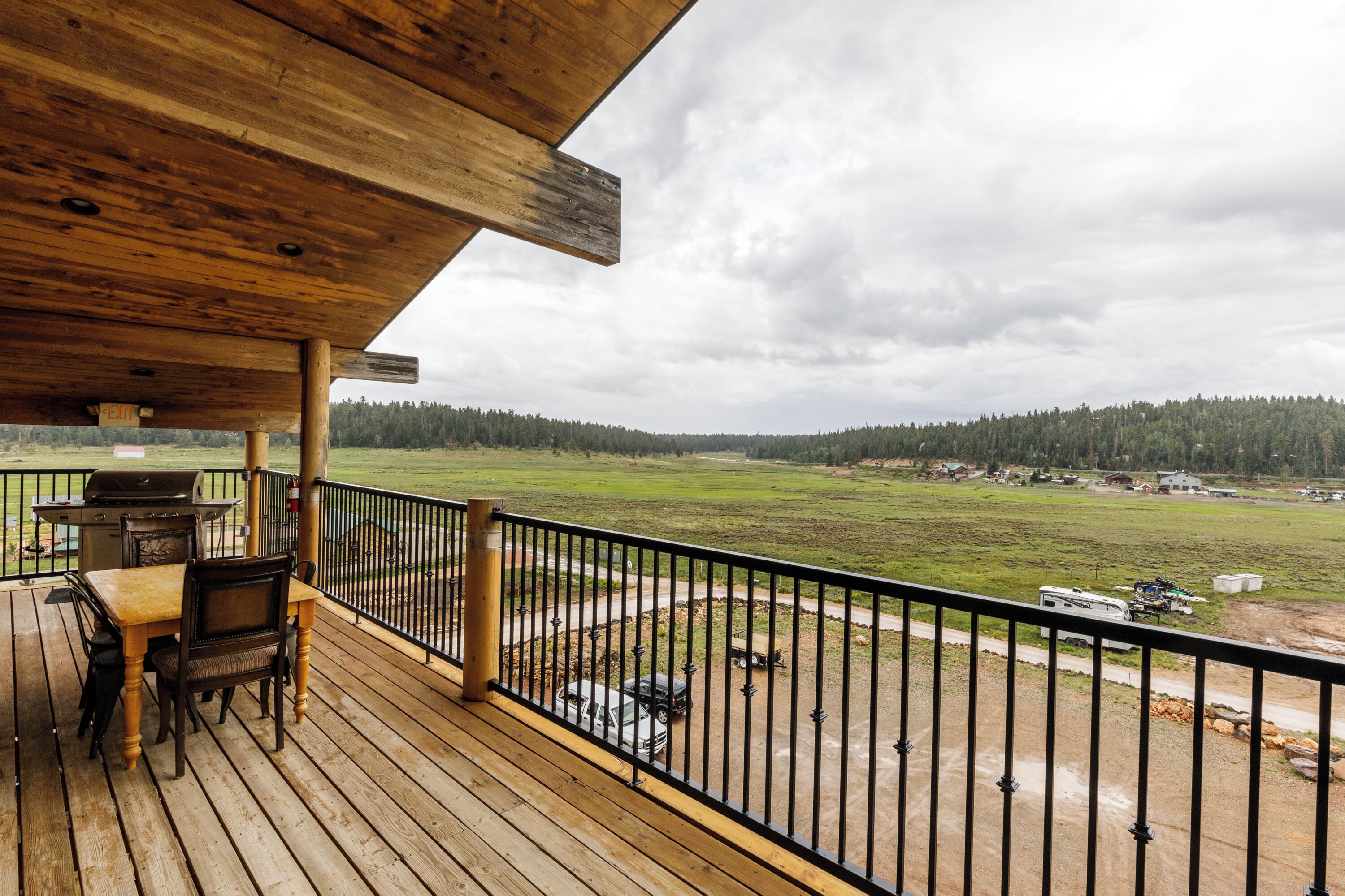 A wooden deck with a grill and chairs, overlooking a grassy field and distant buildings.