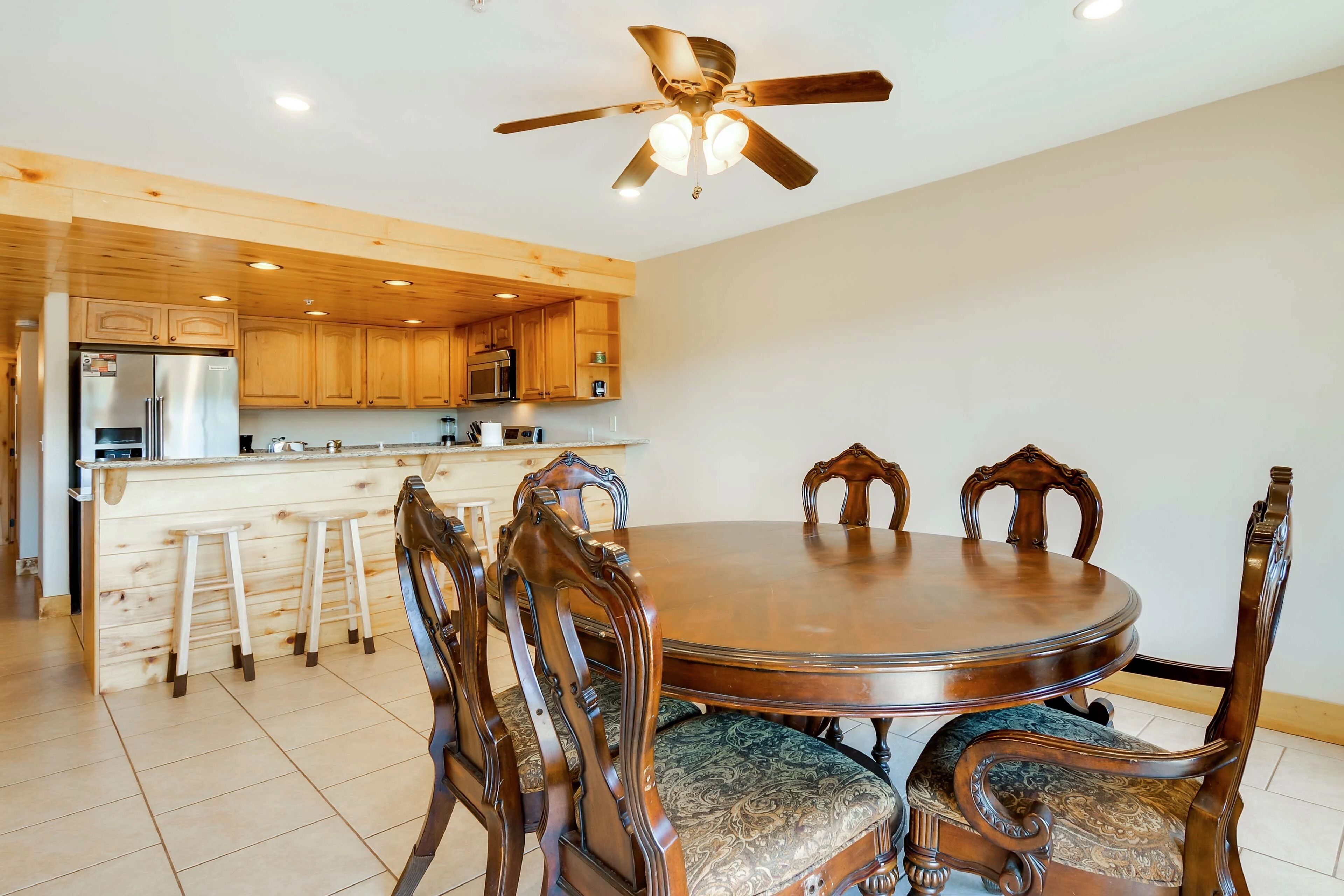 A dining area with a round wooden table and ornate chairs, a kitchen in the background, and a ceiling fan.