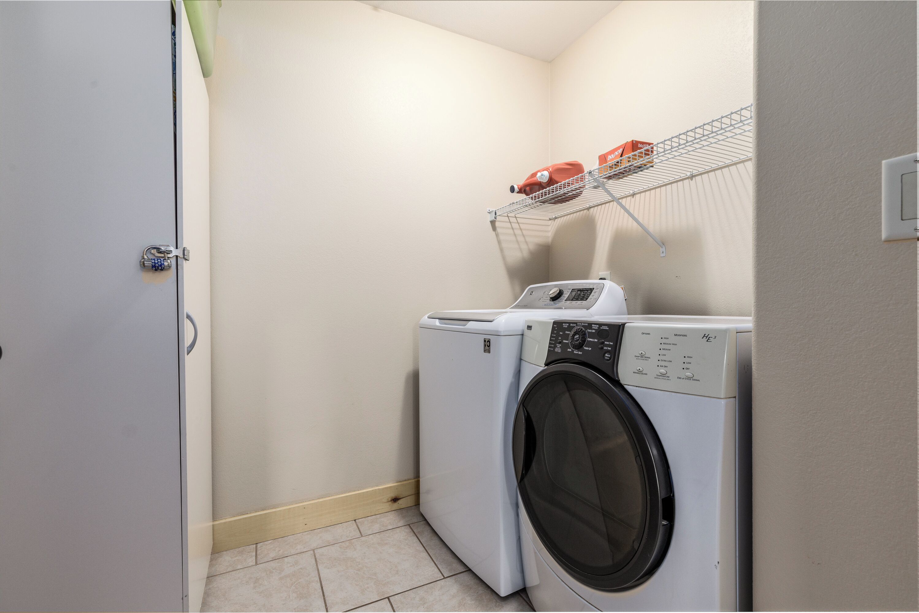 A laundry room with a front-loading washing machine and dryer, a metal shelf with a red item, and a door with a padlock.