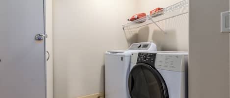 A laundry room with a front-loading washing machine and dryer, a metal shelf with a red item, and a door with a padlock.