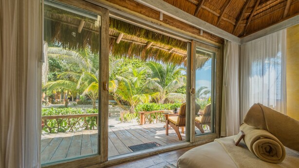 Une chambre avec un lit, une fenêtre donnant sur un jardin tropical et une terrasse en bois à l’extérieur.