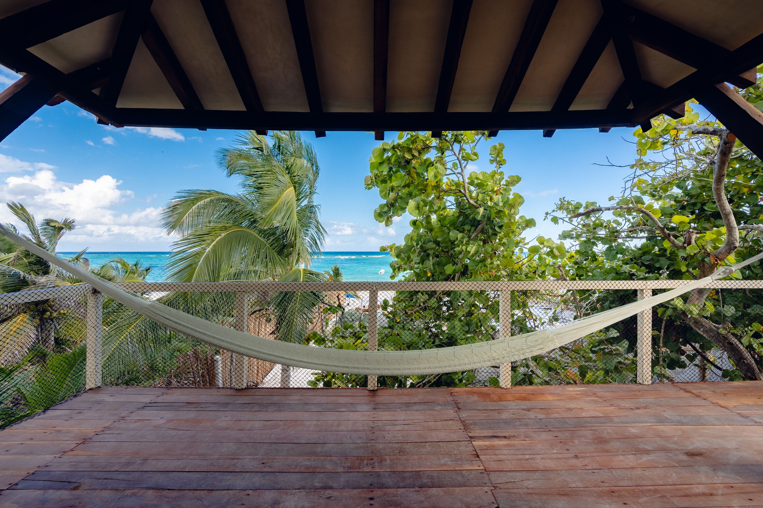 A wooden deck with a hammock, overlooking a beach with palm trees and the ocean.
