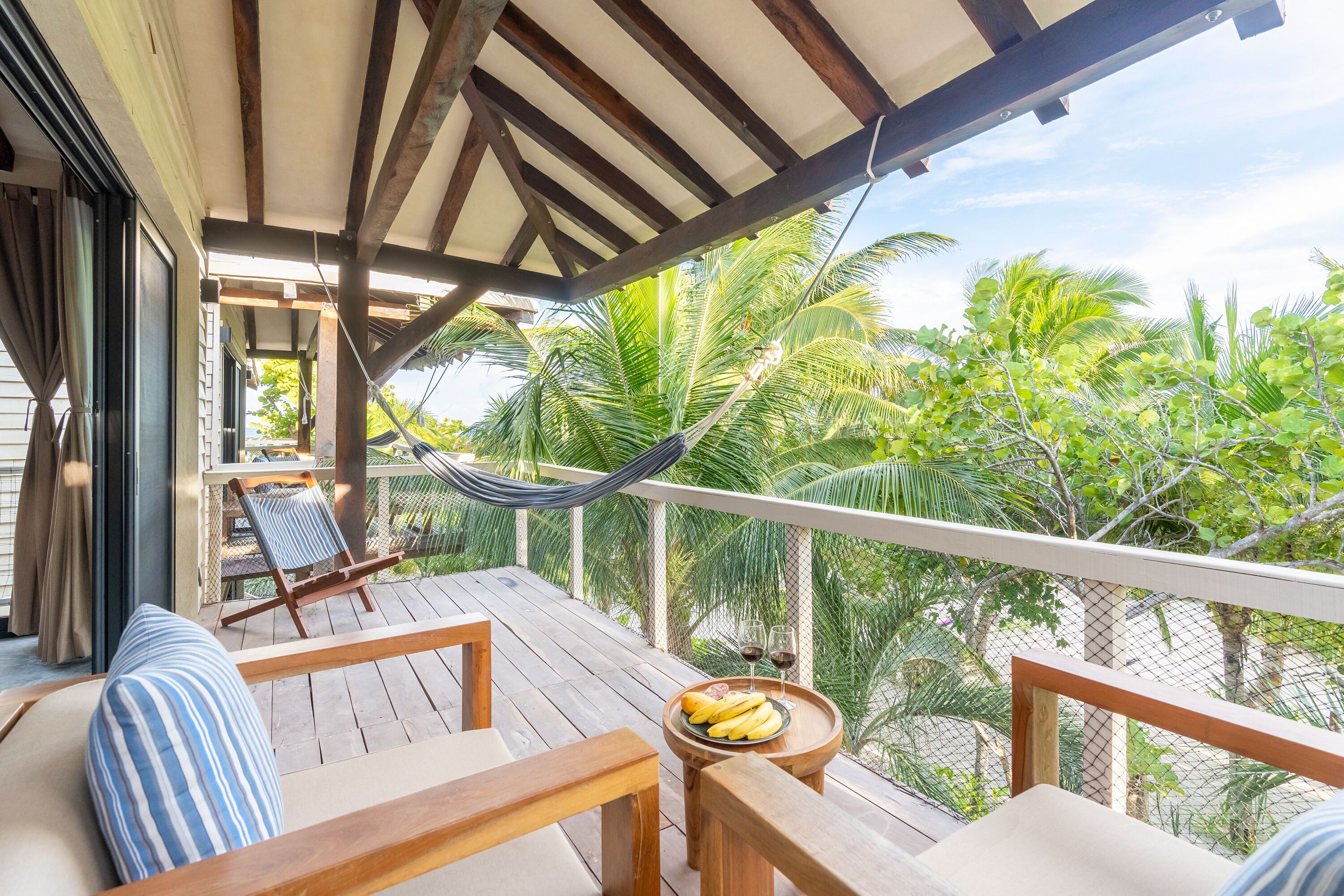 A wooden deck with a hammock, lounge chairs, and a table with a fruit bowl and glasses.