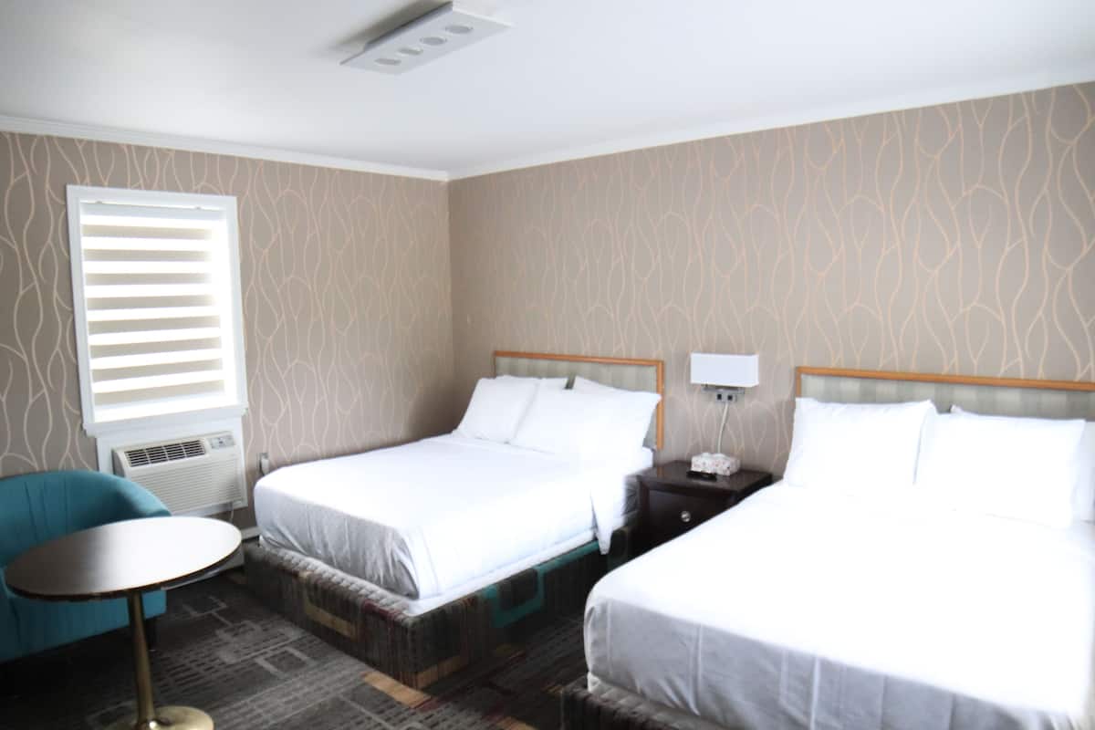 Ein Hotelzimmer mit zwei Betten, einem kleinen runden Tisch, einem Sessel und einem Fenster mit Jalousien.