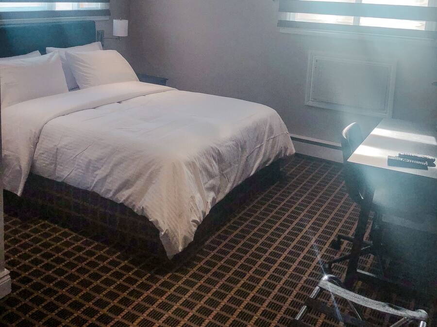 A hotel room with a large bed, a desk with a chair, and a window with blinds.