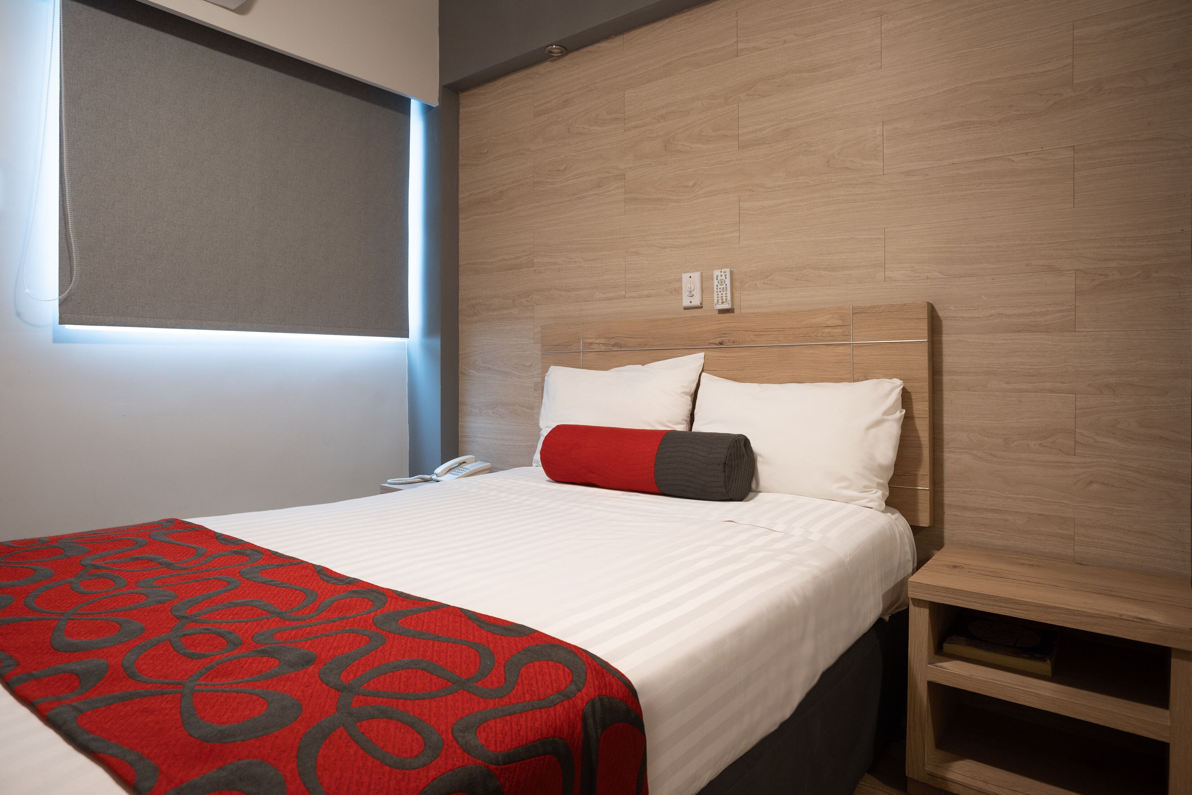A neatly made bed with a red and black patterned blanket, a wooden headboard, and a bedside table with a drawer.