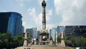 Un monumento con una estatua de un ángel en la cima, rodeado de escalones y estatuas, con un horizonte urbano de edificios modernos de fondo.