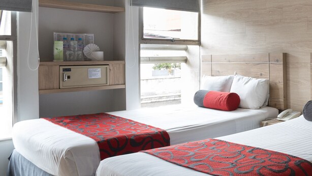 Una habitación de hotel con dos camas, un ventilador de techo, una ventana con cortinas, una mesita de noche y un mueble bajo.