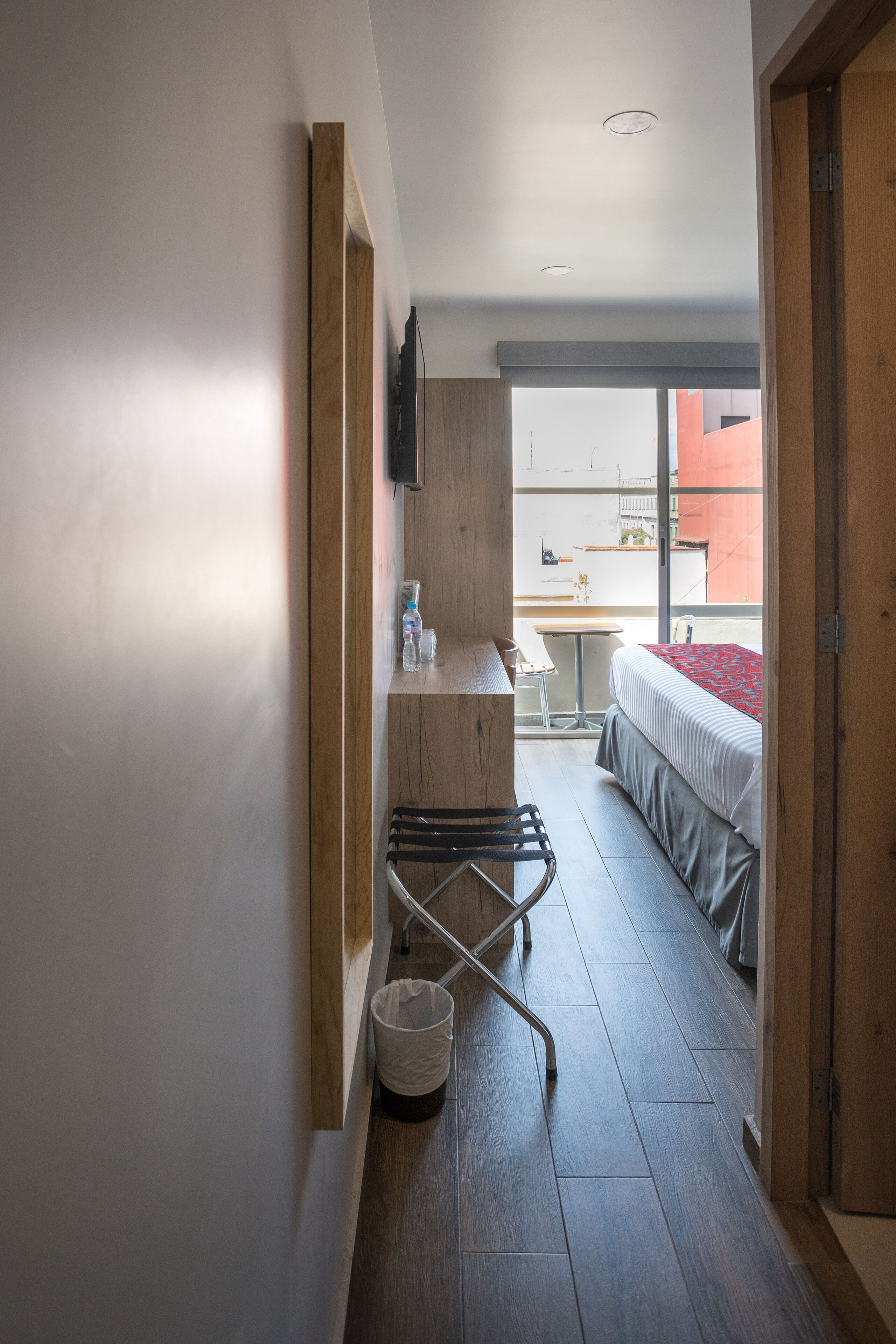 A narrow hallway leading to a bedroom with a bed, a desk, and a chair. There is a small trash bin and a coat rack.