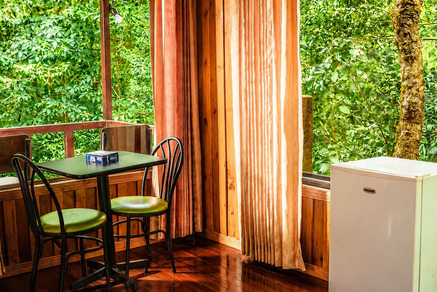 A corner room with a table and chairs, a window with curtains, and a small refrigerator.