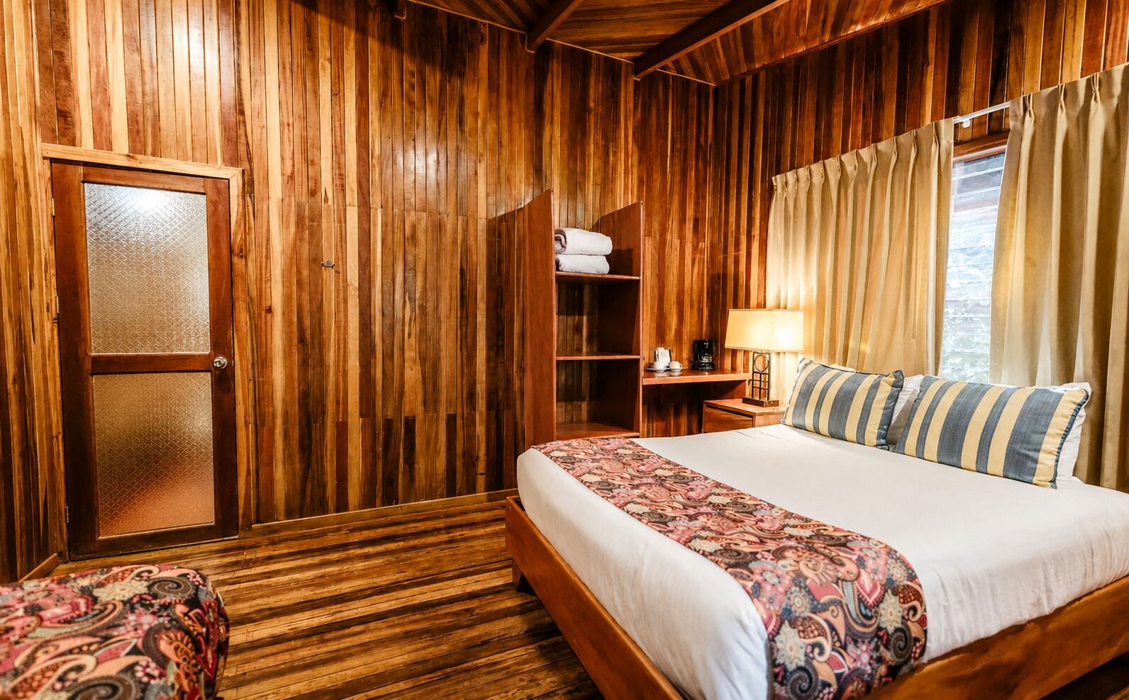 Ein Zimmer mit Holzwänden, einem Bett, einem Sessel, einem Regal mit Handtüchern und einem Fenster mit Vorhängen.