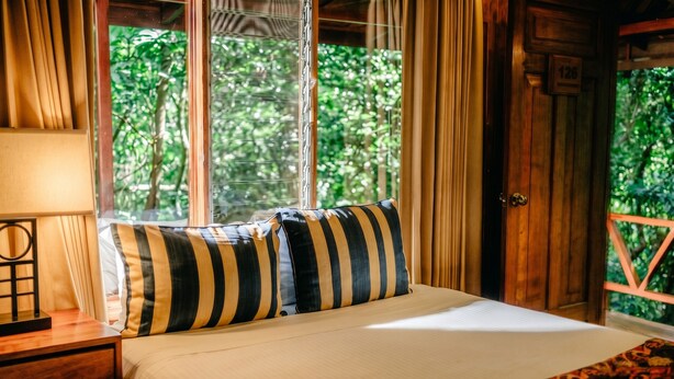 Un lit avec des oreillers à rayures, une table de chevet en bois, une lampe et une fenêtre avec des rideaux.