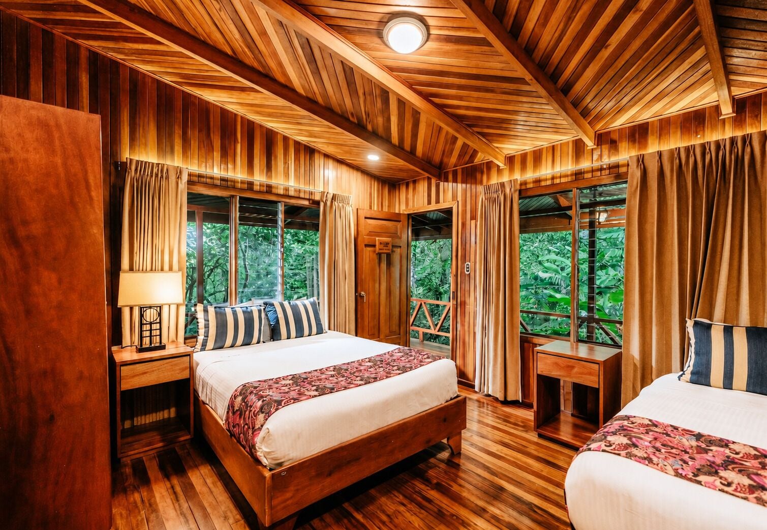Una habitación con paneles de madera, dos camas, una mesita de noche con lámpara y una ventana con cortinas.