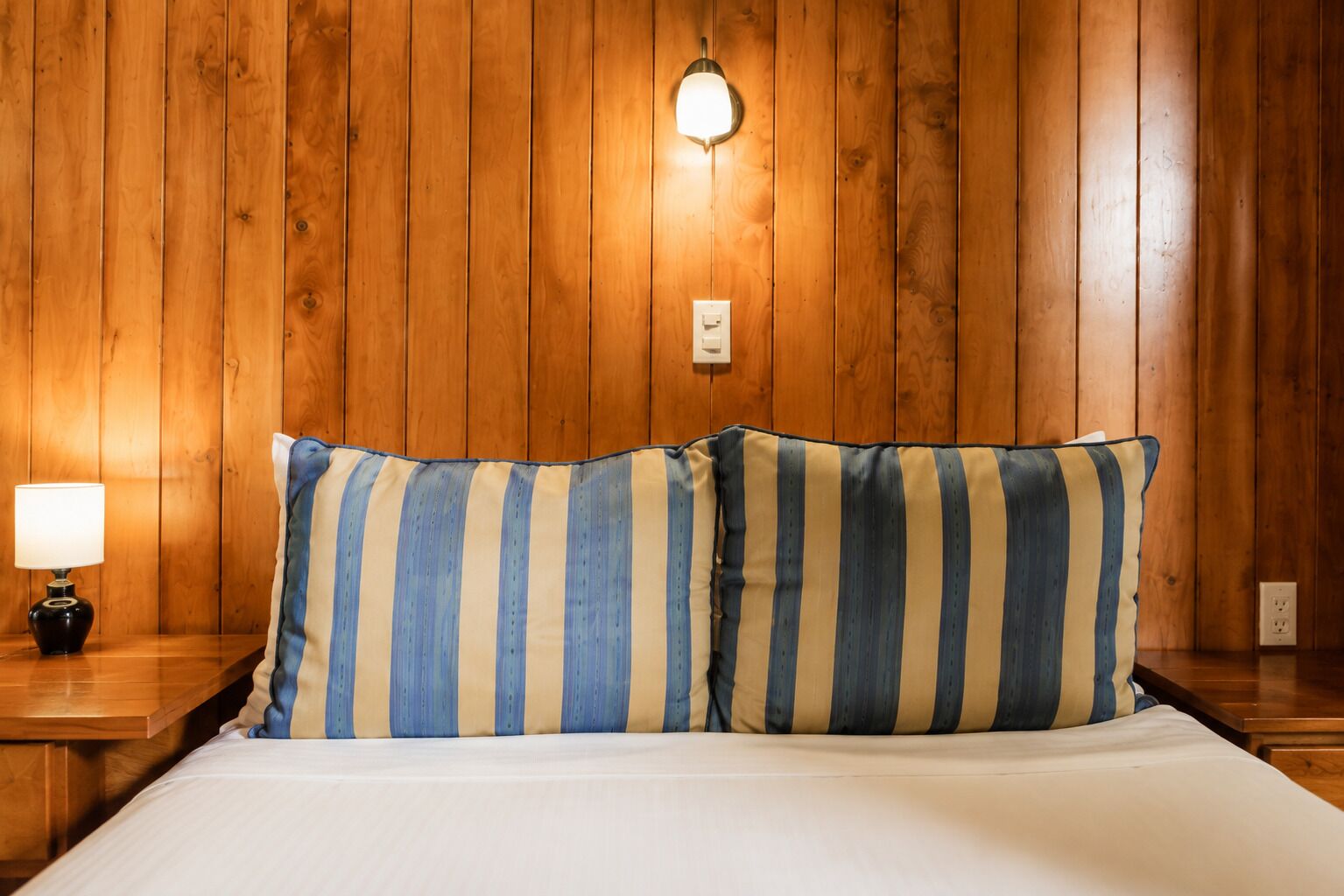 Una habitación con paredes de madera, una cama con ropa blanca, dos almohadas a rayas, una lámpara de noche y un aplique en la pared.