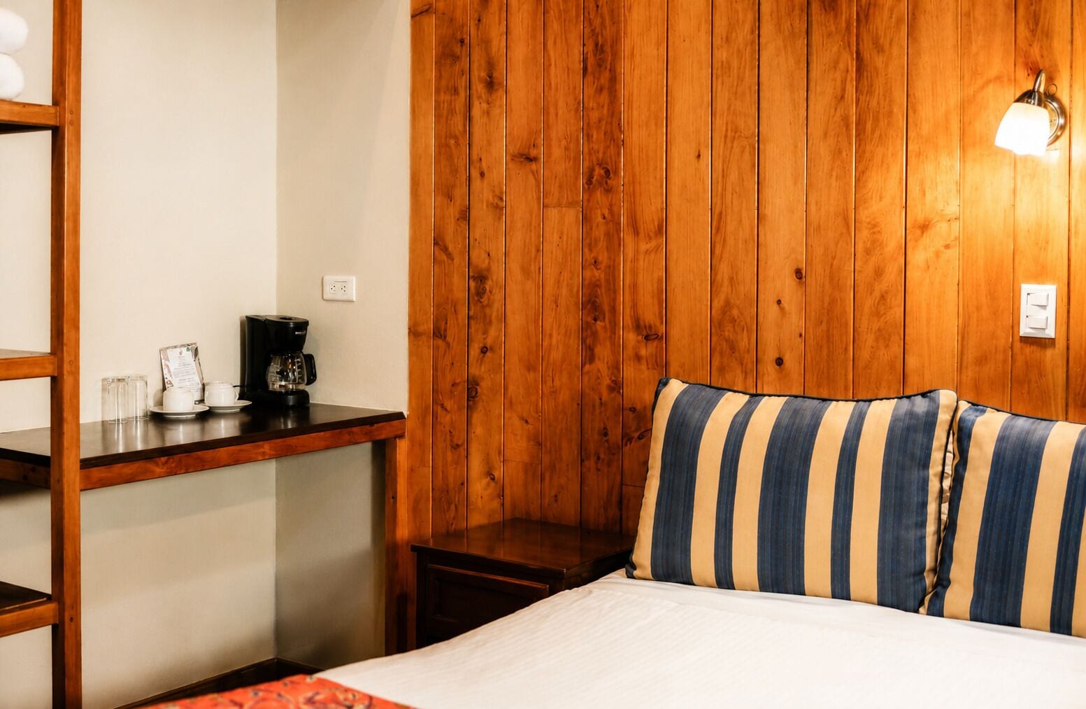 A room with a wooden wall, a bed with striped pillows, a small table with a coffee maker, and a shelf with glasses and a menu.