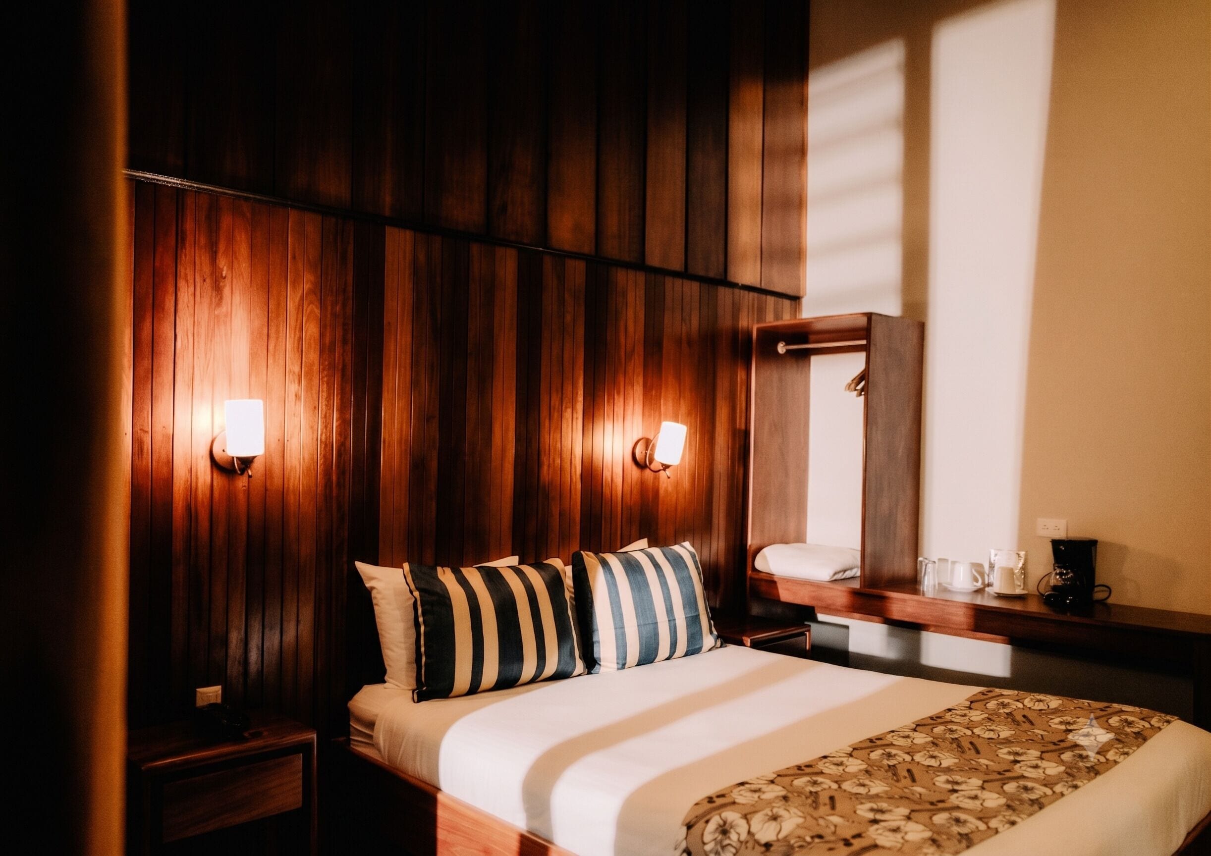 A hotel room with a wooden headboard, a bed with striped pillows, a nightstand, a bedside table with a lamp, and a window with a view of the outside.
