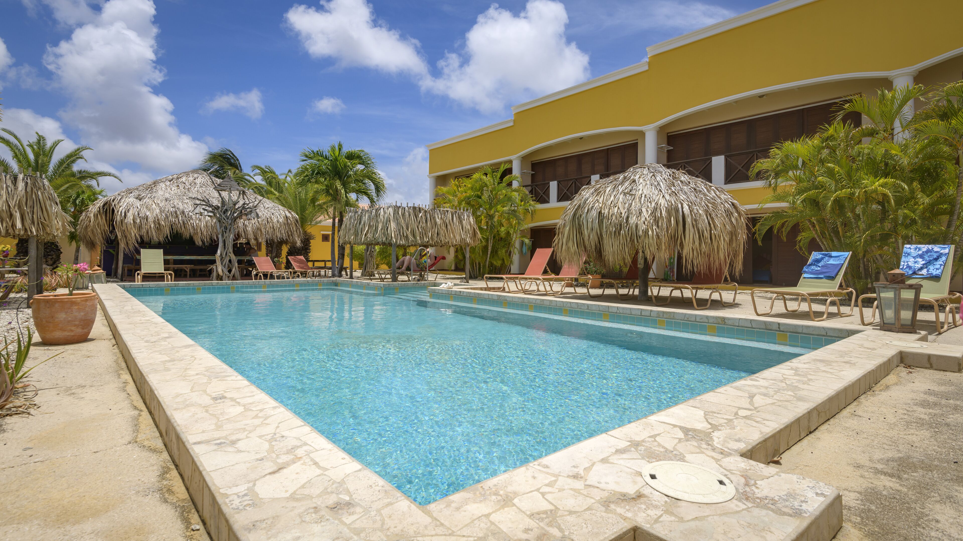 A pool area with lounge chairs and thatched umbrellas, surrounded by palm trees and a yellow building.