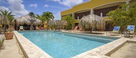 A pool area with lounge chairs and thatched umbrellas, surrounded by palm trees and a yellow building.