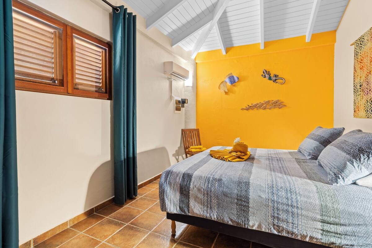 Ein Schlafzimmer mit Bett, einem Fenster mit Jalousien, einem Sessel und einer Wand-Klimaanlage.