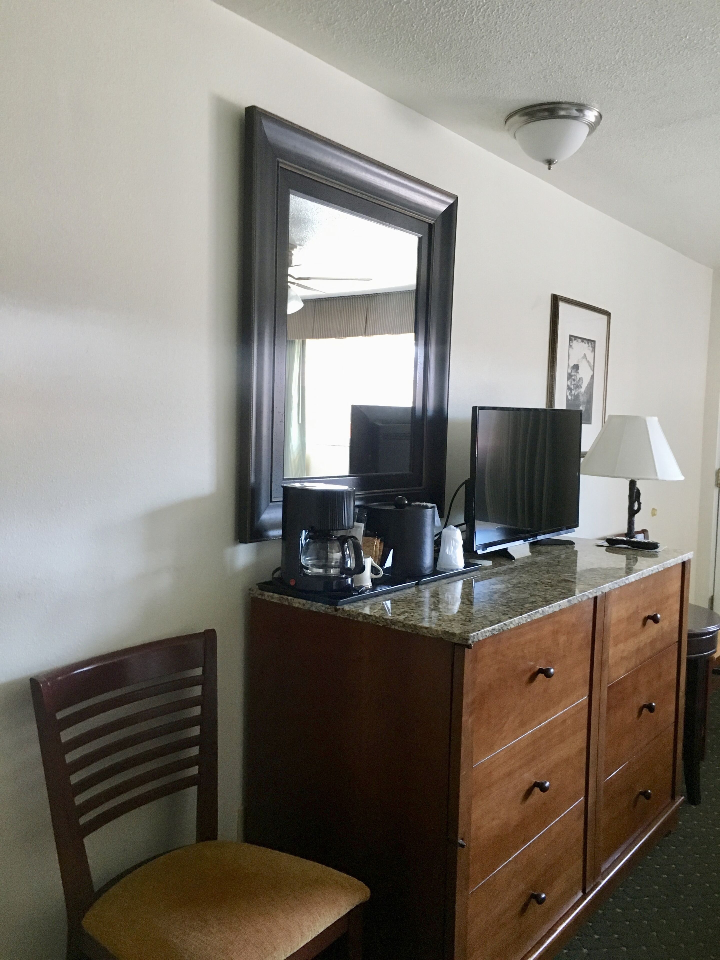 Una habitación de hotel con un aparador de madera, televisor, cafetera, una silla y una lámpara.