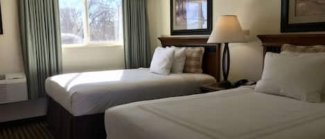 A hotel room with two single beds, a bedside table with a lamp, a window with curtains, and a framed picture on the wall.