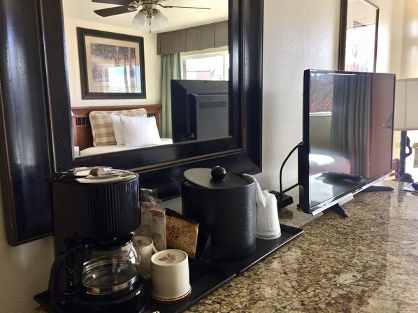 A hotel room with a large mirror, a coffee maker, a kettle, a remote control, and a small table with a cup and a tissue box.