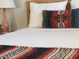 Una cama con un almohadón blanco y un almohadón estampado, una colcha blanca y una manta a rayas rojas y negras.