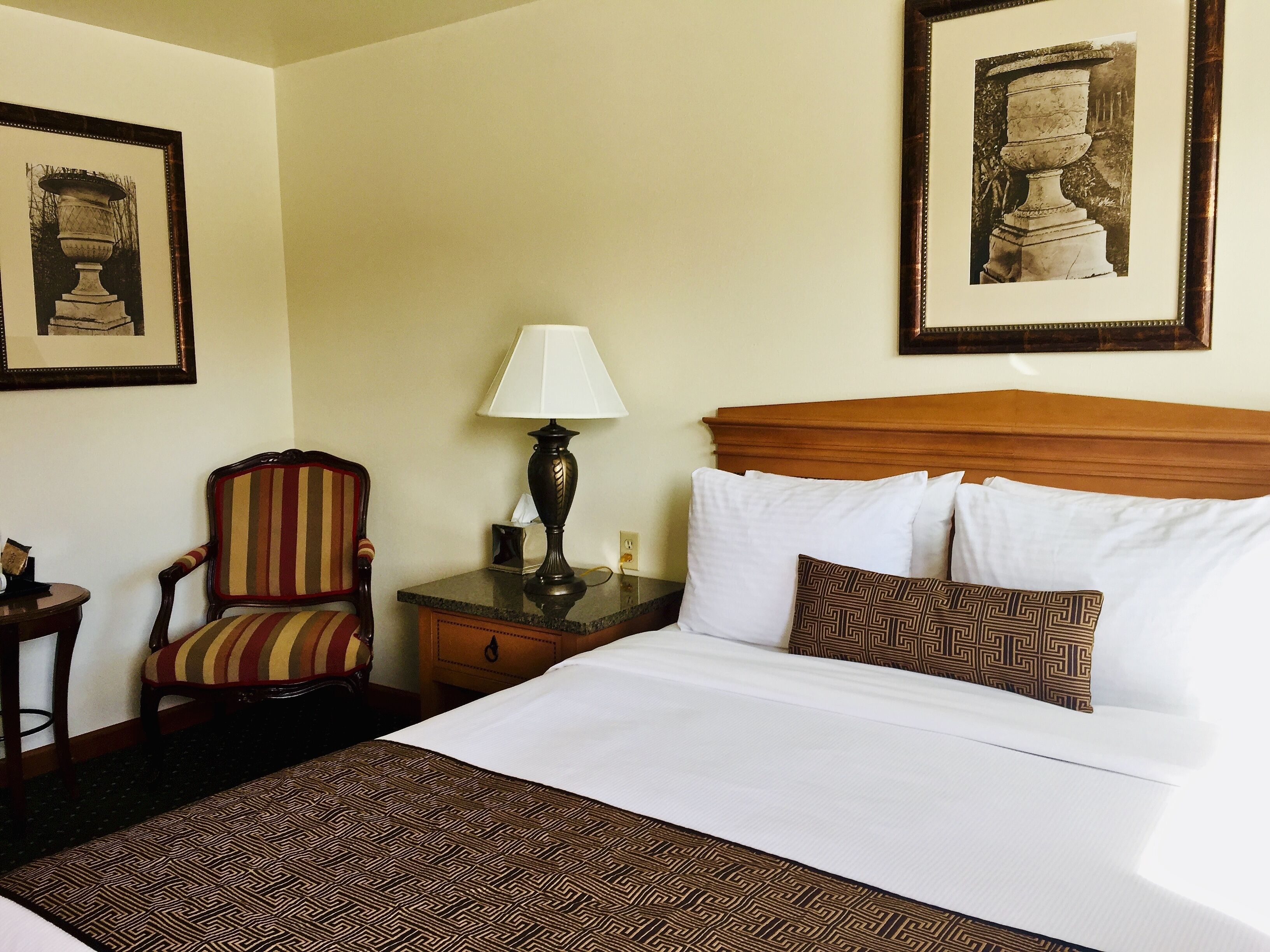 Una habitación de hotel con una cama, una silla, una mesita de noche, una lámpara y dos cuadros en la pared.