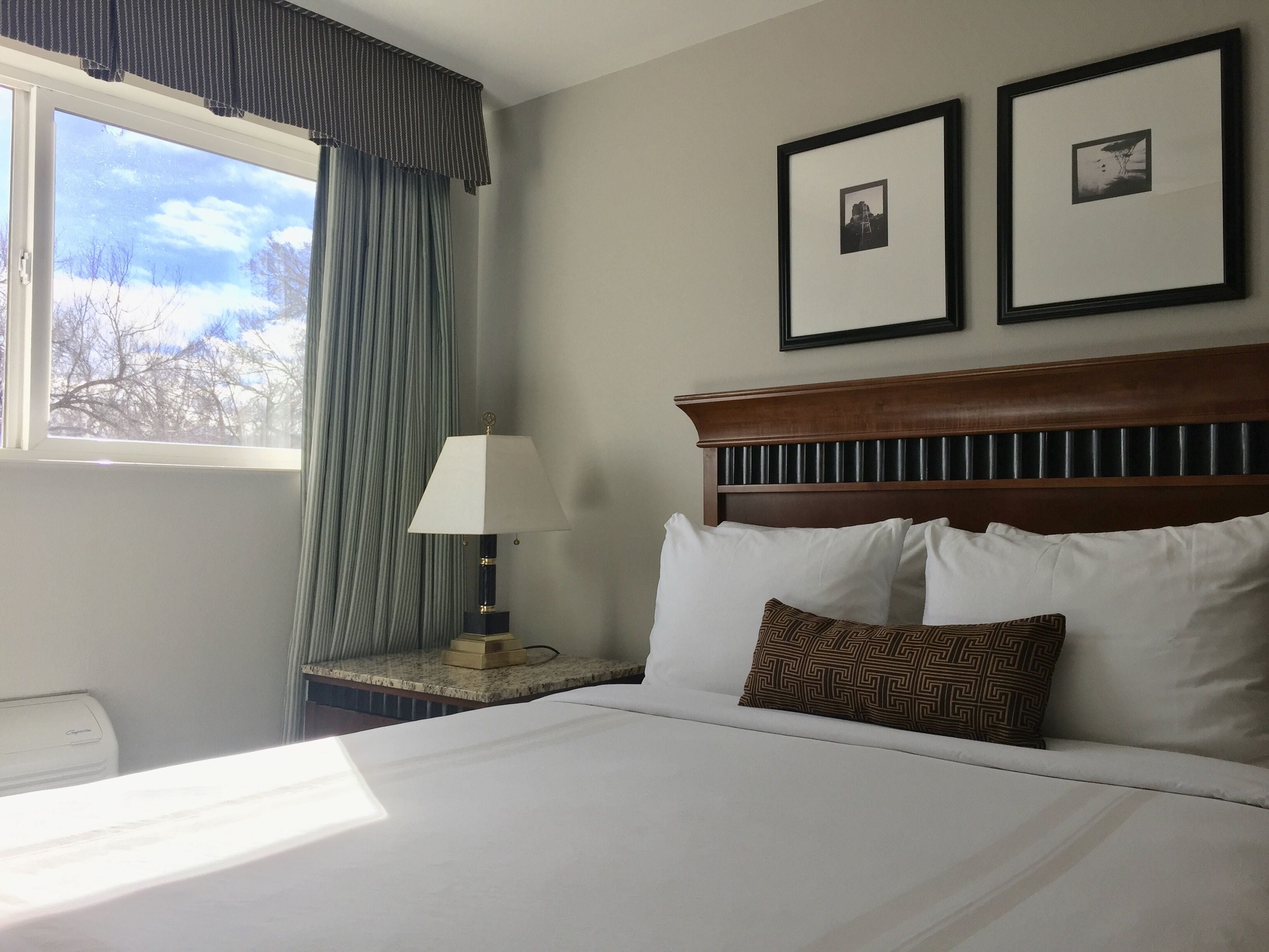 A hotel room with a large bed, a bedside table with a lamp, two framed pictures on the wall, and a window with curtains.