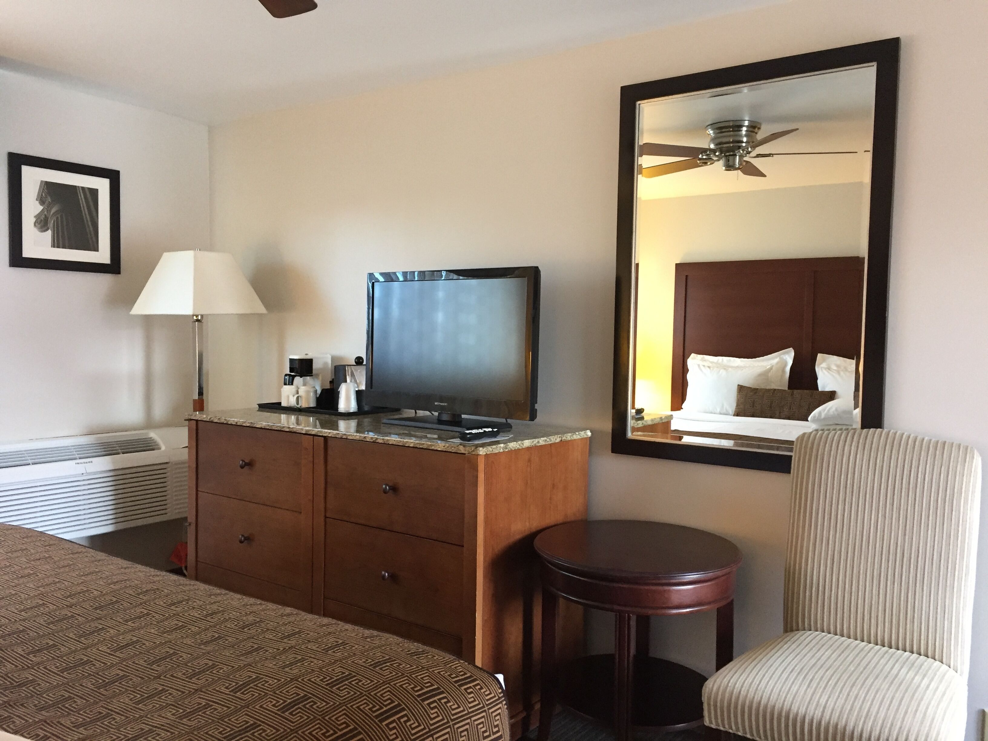Quarto de hotel com uma cama, cômoda de madeira com TV de tela plana, abajur, mesa redonda pequena e um espelho grande refletindo o quarto.