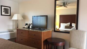 Quarto de hotel com uma cama, cômoda de madeira com TV de tela plana, abajur, mesa redonda pequena e um espelho grande refletindo o quarto.