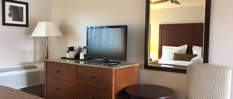 A hotel room with a bed, a wooden dresser with a flat-screen TV, a lamp, a small round table, and a large mirror reflecting the room.