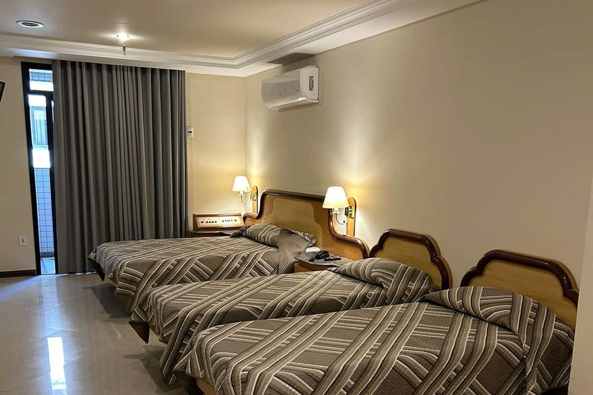 Habitación de hotel con tres camas, un ventanal grande con cortinas y piso de baldosas.