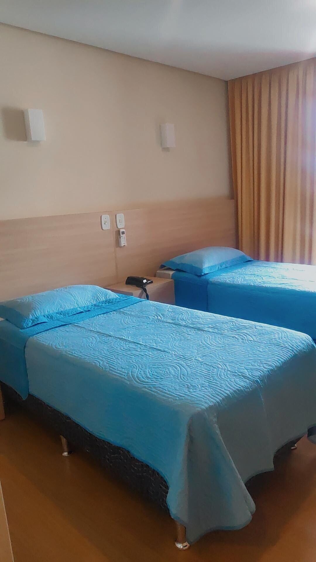 Een hotelkamer met twee bedden, elk met een blauwe spreideken, een houten hoofdeinde, een nachtkastje met een telefoon en een gordijn voor het raam.